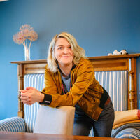 Eine Frau mit blonden, langen Haaren in einem goldfarbenen Blouson lehnt sich lässig über einen Stuhl. Hinter ihr ein blau-weiß gestreiftes Biedermeier-Sofa vor einer blauen Wand, eine Gitarre in einer Ecke. Eine Snoopy-Figur liegt auf der Sofalehne, ein paar Trockenblumen in einer Vase, ein paar zusammengelegte Decken. Ein Fenster mit blauem Vorhang. Vor dem Stuhl steht ein antiker Tisch. Aufmerksam und leicht lächelnd blickt die Frau in Richtung des Fotografen.