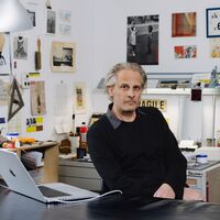 Ein Mann sitzt an einem Schreibtisch vor einem Laptop und sieht in die Kamera. Dahinter herrscht das kreative Chaos: Auf Tischen und in Regalen sind viele verschiedene Gegenstände zu sehen – Stifte, Kleber, kleine Geräte und Aufbewahrungsboxen. An den Wänden hängen Zeichnungen, Fotos, Hinweis- und Gefahrenschilder und vieles mehr.