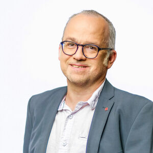 Portrait eines grauhaarigen Herrn mit Brille in einem weißen Hemd und grauem Jackett.