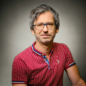 Portrait eines grauhaarigen Herrn mit Brille, Dreitagebart und rotem, gemusterten Poloshirt.