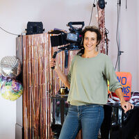 Nathalie Wiedemann Eine Frau steht mit einer Filmkamera auf der Schulter in einer kunterbunten Kulisse. Im Vordergrund ein Flügel, der mit bunten Schirmmützen bepackt ist; ein kupferfarben schillernder Paravent; Diskokugeln. Durch einen Türbogen hindurch ist eine bunte Küche und eine mit Goldpapier gedeckte Tafel mit Partyutensilien zu sehen. An der Wand das Wort "Happy" in bunten Buchstaben.