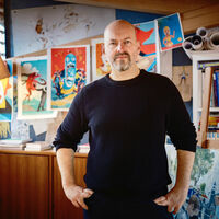 David von Bassewitz Ein Mann steht in der Mitte eines Raumes, die Hände in die Seiten gestemmt, und guckt in die Kamera. Hinter ihm hängen aufgereiht bunte Illustrationen, ein Regal mit Büchern, einige Papierrollen und ein großes Bild vor einer Staffelei.