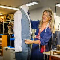 Mirjam Muschel In einer Kostümwerkstatt steckt eine Frau im blauen Kleid und langem blonden Haar einen Herrenanzug auf einer Schneiderpuppe fest. Er besteht aus Hose und Weste über einem weißem Hemd mit Fliege. Das Sakko liegt auf einem Tisch neben einem Bügeleisen.